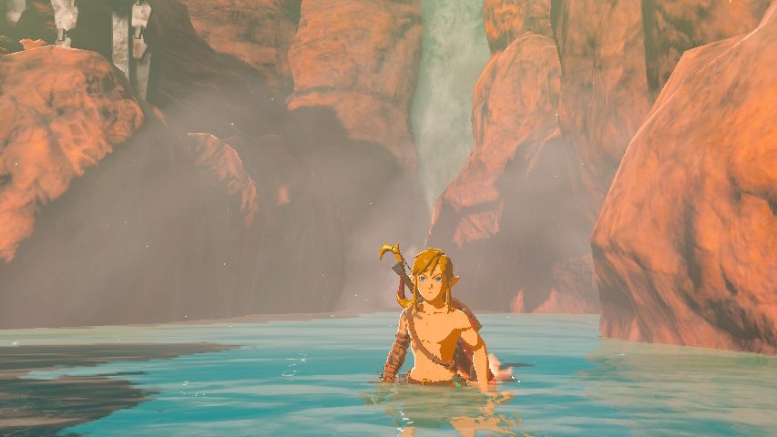 Link is not wearing a shirt, and he stands submerged in a turquoise hot spring which steam rising from it. The spring is surrounding by red rock formations.